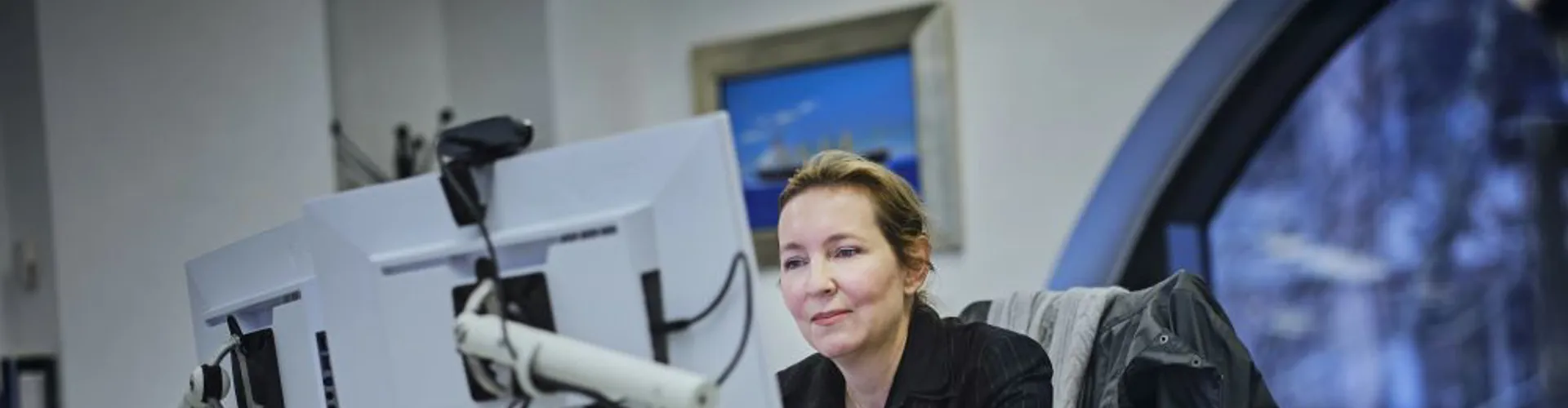 Woman in a black jacket seated at a modern desk focused on two computer monitors.
