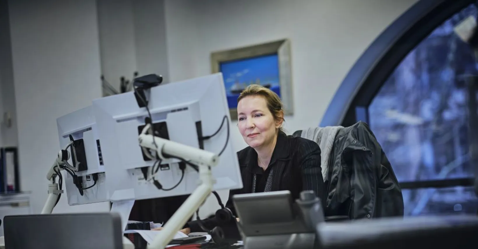 Woman in a black jacket seated at a modern desk focused on two computer monitors.