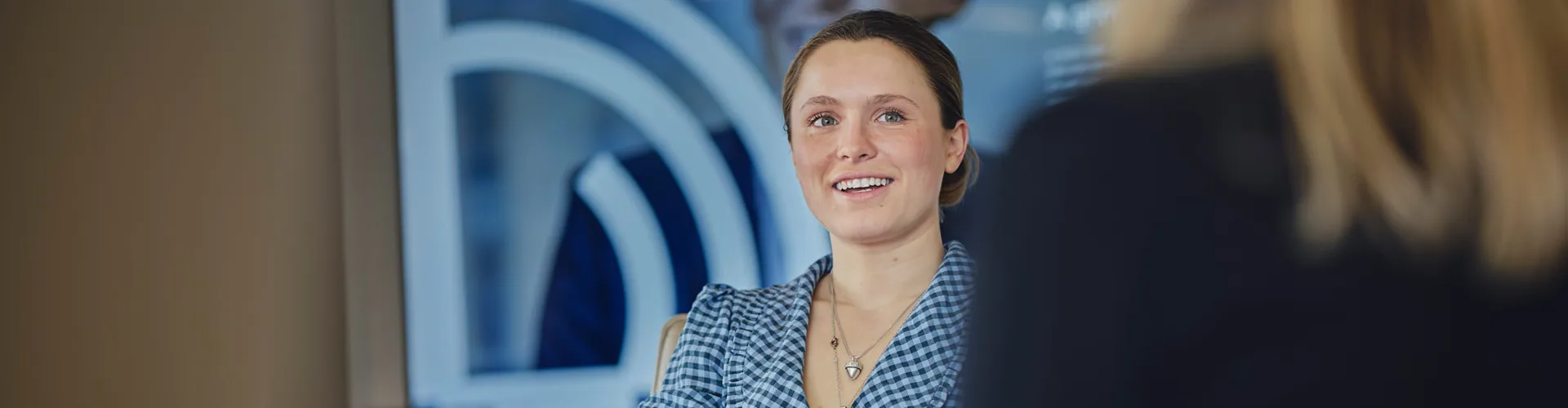 Smiling young woman with light brown hair in a checkered blouse seated at a table.
