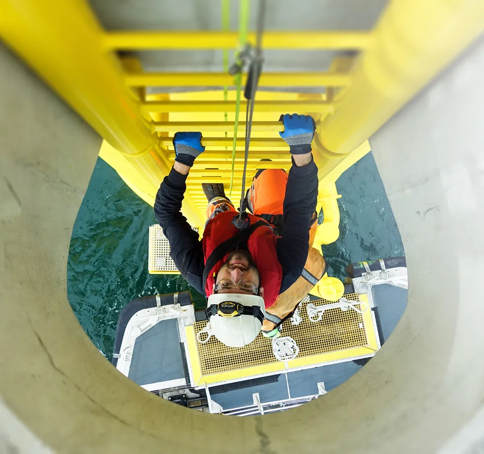Manual high worker offshore climbing on wind-turbine on ladder and transfer vessel waiting under wind turbine