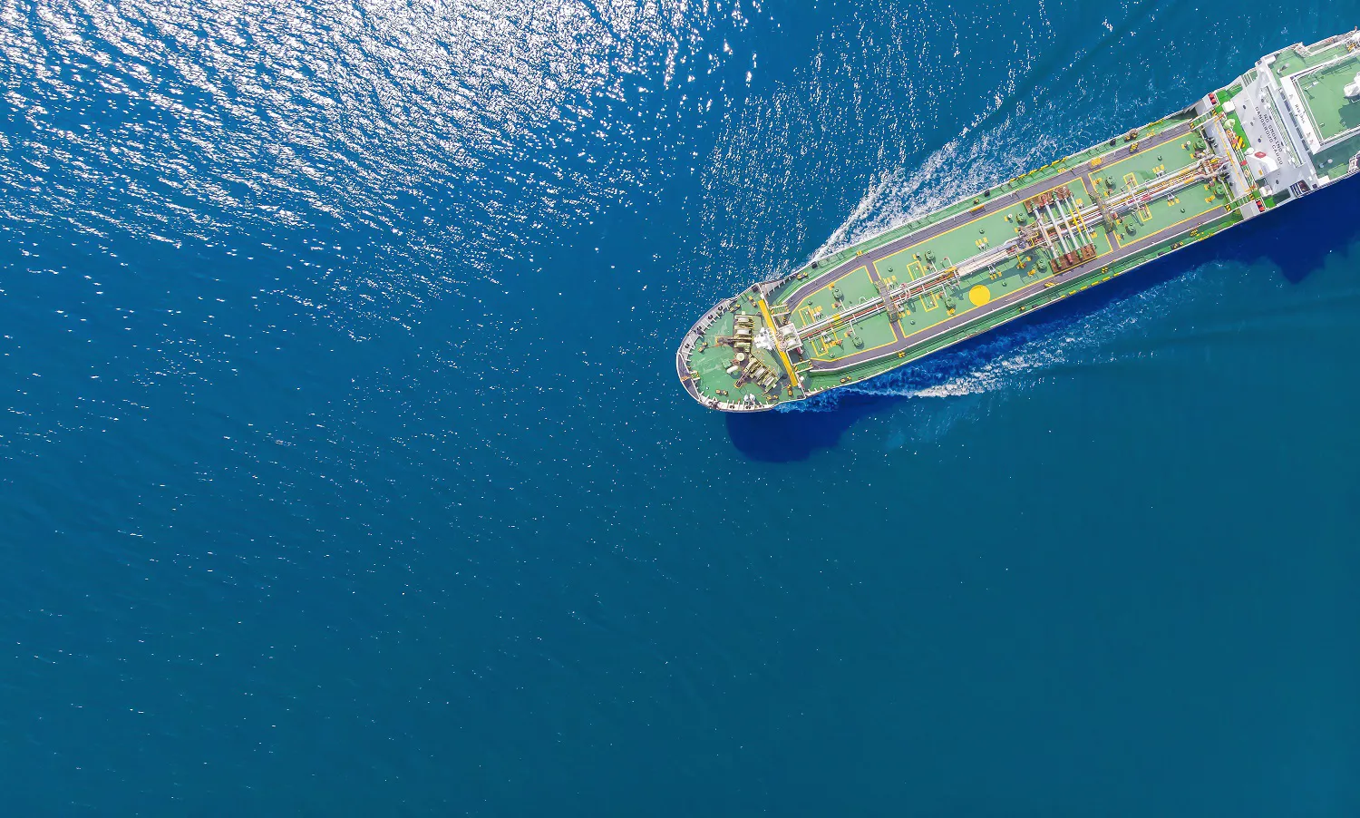 Aerial view of a large ship navigating through deep blue water, with sunlight reflecting off the surface.