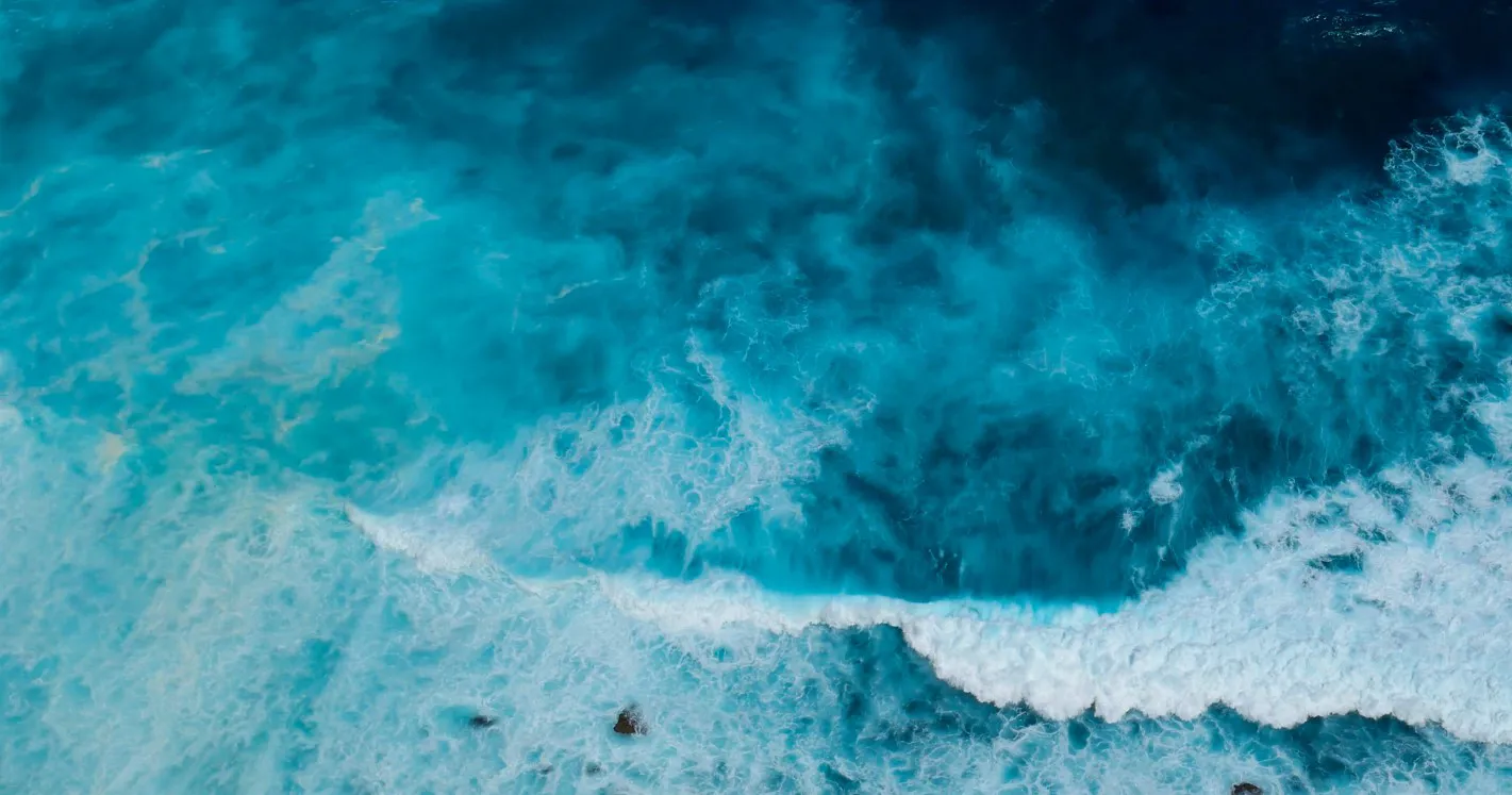 Aerial view of turquoise ocean with white waves crashing near rocky shore.