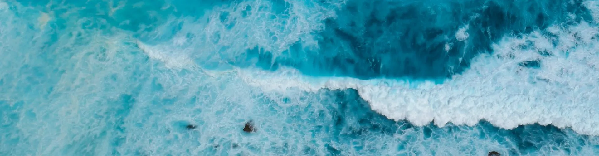 Aerial view of turquoise ocean with white waves crashing near rocky shore.