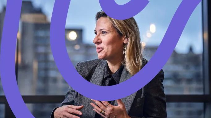 Woman in a blazer speaking, city backdrop visible.