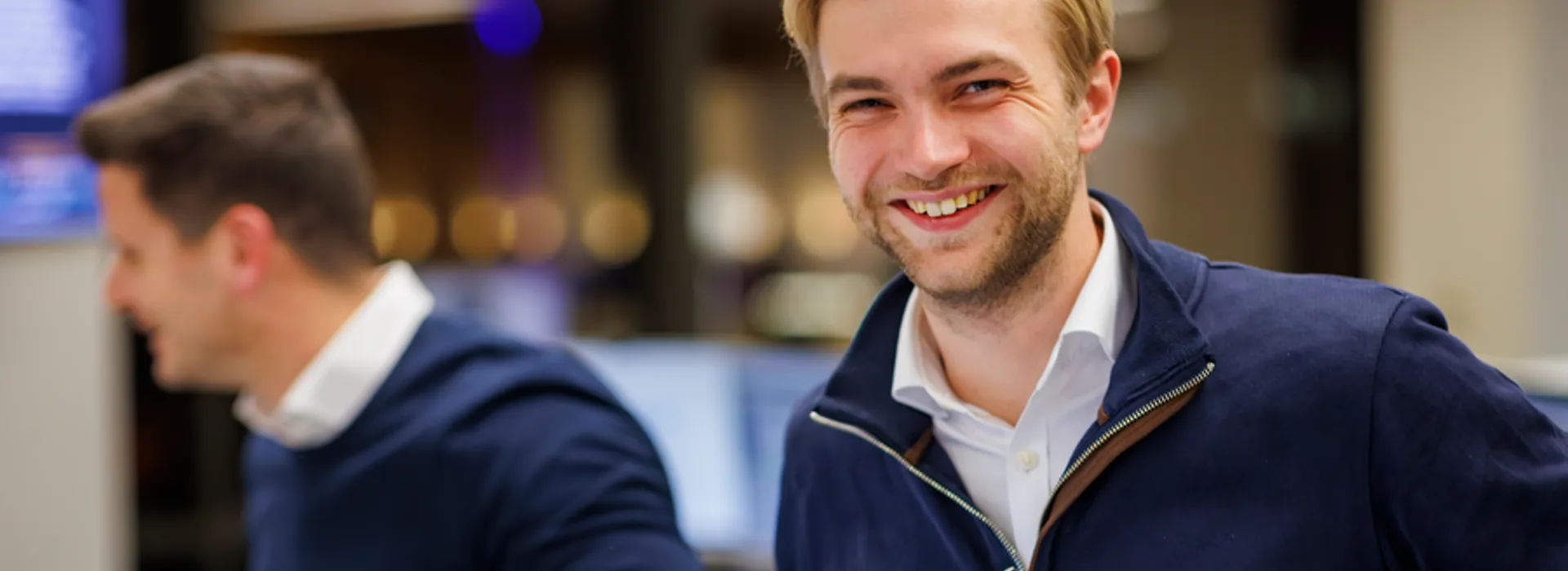 Smiling young man with blonde hair and beard in a navy sweater, with colleagues and monitors blurred behind.
