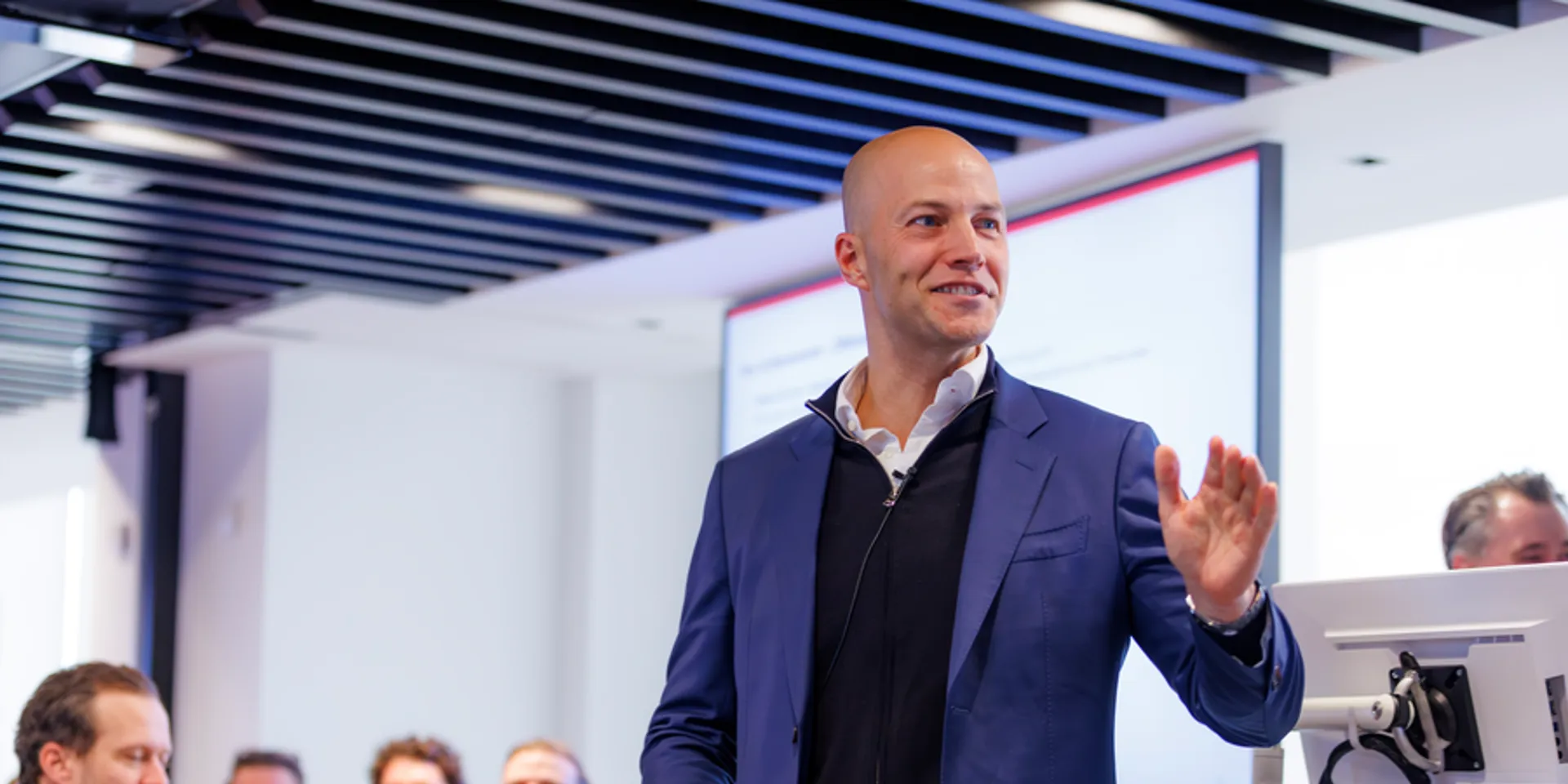 Smiling man in a blue blazer speaking and gesturing to a seated audience.