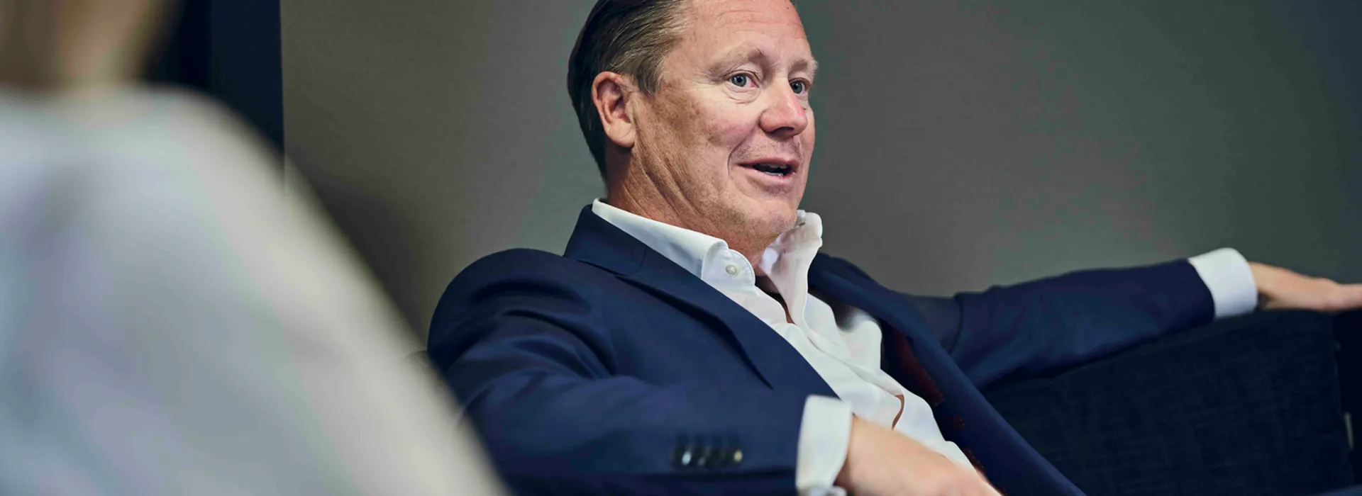 Man in a dark suit seated on a couch, engaging in conversation.