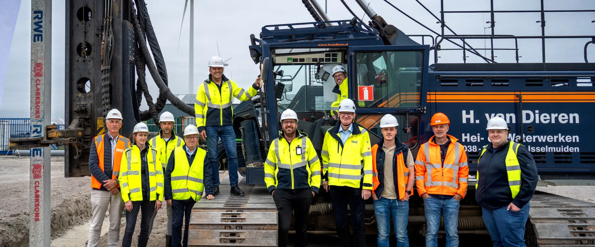 Group of 11 in safety gear posing by heavy machinery with a wind turbine backdrop.