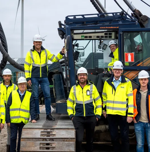 Group of 11 in safety gear posing by heavy machinery with a wind turbine backdrop.
